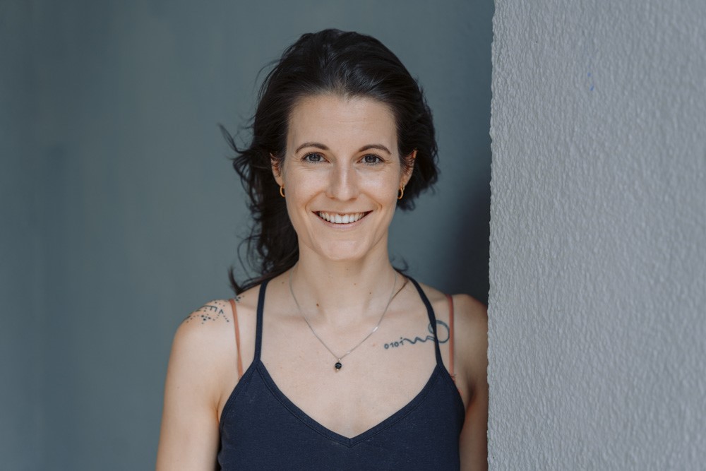 White woman in dark tank top, white smile, dark hair, tattoos. Narrow, gray wall to the right and gray background.
